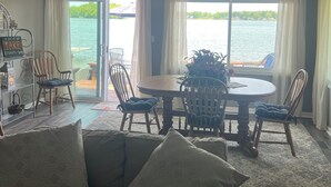 Een woonkamer met een bank, een eettafel met stoelen, een plafondventilator en uitzicht op een wateroppervlak door grote ramen.