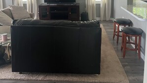 Een woonkamer met een grote zwarte bank, een flatscreen televisie op een houten kast, een klok aan de muur en een keuken met krukjes.