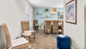A dining area with wicker chairs and a table, a blue ottoman, and a hanging lamp.