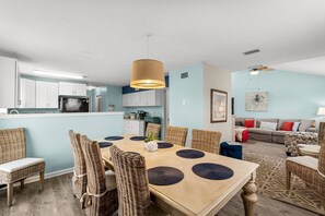 Eine moderne Küche mit weißen Schränken, ein Essbereich mit rundem Tisch und Korbstühlen sowie ein Wohnzimmer mit Sofa und gemustertem Teppich.
