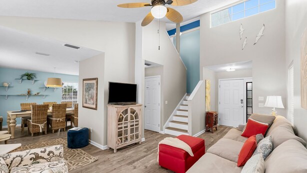 A spacious living room with a large sofa, a dining area with wicker chairs, a TV on a wooden cabinet, and a staircase leading to another level.