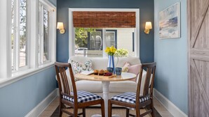 Une pièce chaleureuse avec un mur bleu, un canapé blanc, deux chaises en bois, une petite table ronde ornée d’un vase de fleurs et une fenêtre avec des stores.