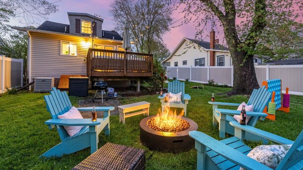 Un jardin avec un foyer entouré de chaises Adirondack bleues, une terrasse en bois et une clôture blanche.