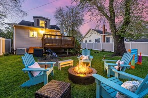 A backyard with a fire pit surrounded by blue Adirondack chairs, a wooden deck, and a white fence. - Charming Beach Cottage Near Cafés & Shops (North Beach)