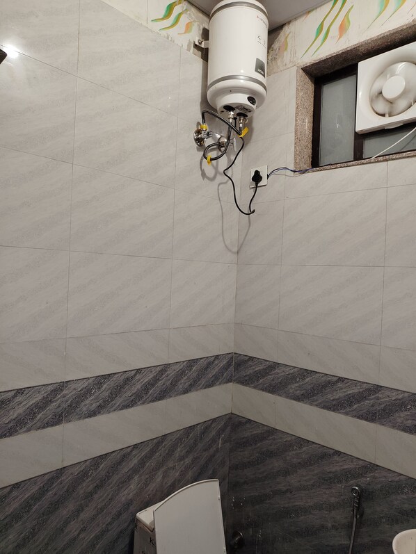 A bathroom with a white water heater mounted on the wall, a shower area with a dark marble floor, and a window with a decorative frame. - Khatu Shyam Niwas (Danta Ramgarh)
