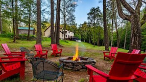 A campfire surrounded by red Adirondack chairs in a forested area with a house in the background. - Couple s Oasis Pool Stream Firepit Steam Room (Saugerties)