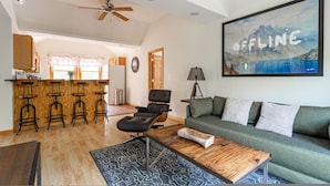 A spacious living area with a green sofa, a wooden coffee table, and a black armchair. There is a kitchen area with wooden bar stools and a ceiling fan. A large painting of a mountain landscape is on the wall. - Couple s Oasis Pool Stream Firepit Steam Room (Saugerties)
