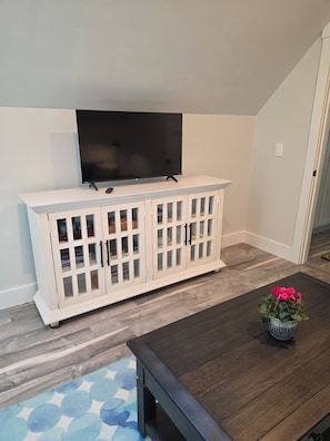 A room with a dark wooden dining table, a white cabinet with glass doors, and a flat-screen TV mounted on the wall. - Round Pond Maine - Charming apartment for 4 with AC, WiFi and ocean view. (Round Pond)
