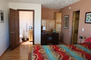 Une chambre à coucher avec un lit en bois, un bureau avec une chaise, une armoire et une salle de bain visible à travers une porte ouverte.