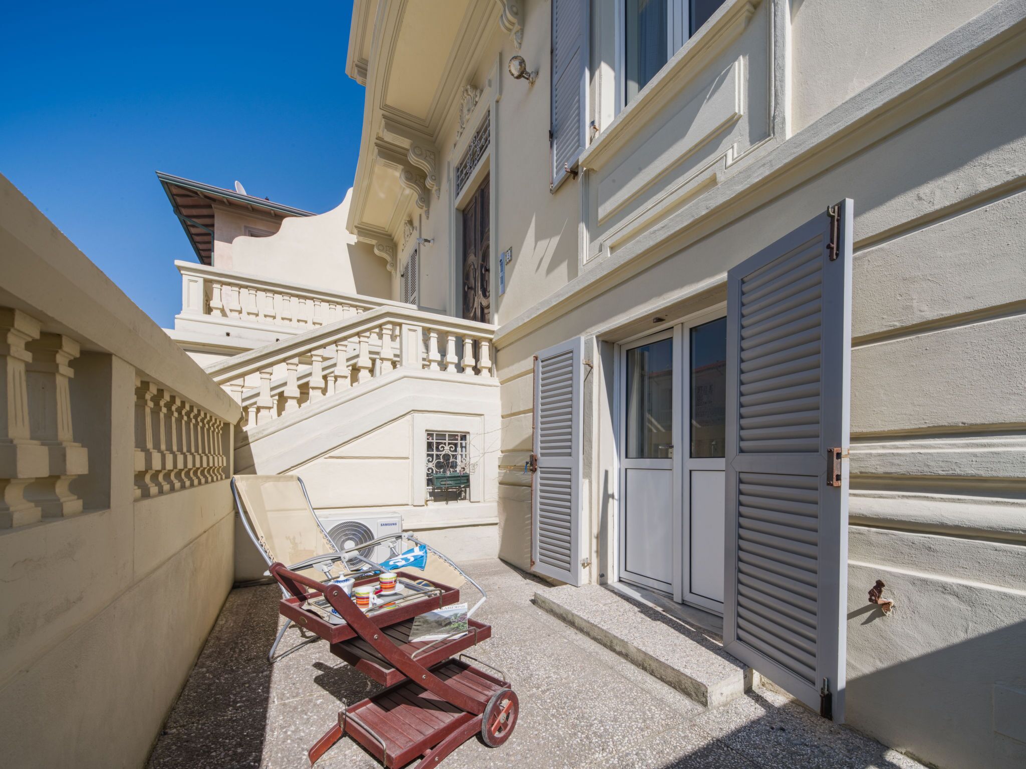 A narrow courtyard with a wooden cart, a Samsung air conditioning unit, and a staircase leading to a balcony.