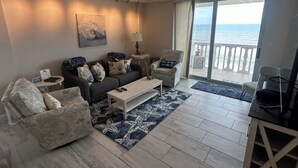 A living room with a sofa, armchairs, a coffee table, and a TV. There is a view of the ocean through a balcony.