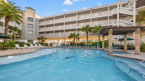Un espace piscine d’hôtel comprenant plusieurs chaises longues, un palmier et un immeuble à plusieurs étages doté de balcons.