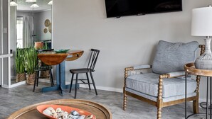 Un salon avec un téléviseur, un canapé gris, une chaise en bois, une table à manger avec des chaises et des plantes en pot.