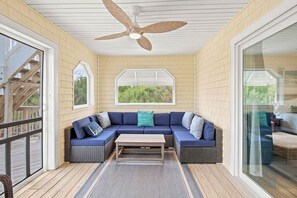A covered outdoor seating area with a ceiling fan, a curved sofa with cushions, and a wooden table.