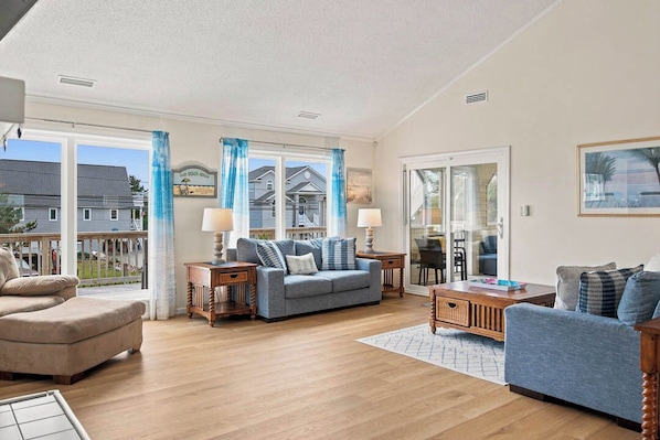 A spacious living room with a blue sofa, beige armchair, and a wooden coffee table. Large windows with blue curtains provide ample natural light.