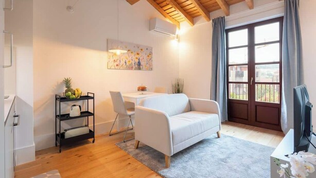 A modern living room with a white sofa, a small desk with a chair, a black shelf with fruits, and a painting on the wall.
