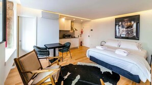 A modern hotel room with a bed, a desk, and a chair. There is a kitchen area in the background.