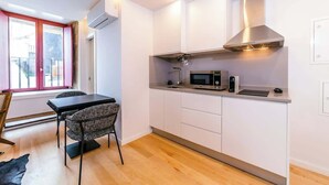 A compact kitchen with white cabinets, a stainless steel range hood, a microwave, and a sink. There is a dining table with two chairs and a window with pink curtains.