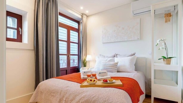 A hotel room with a bed, a breakfast tray, a window with curtains, a white wall with a painting, and a small white shelf with a plant.
