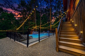 A wooden staircase leading to a pool area surrounded by a black metal fence, with string lights and trees in the background.
