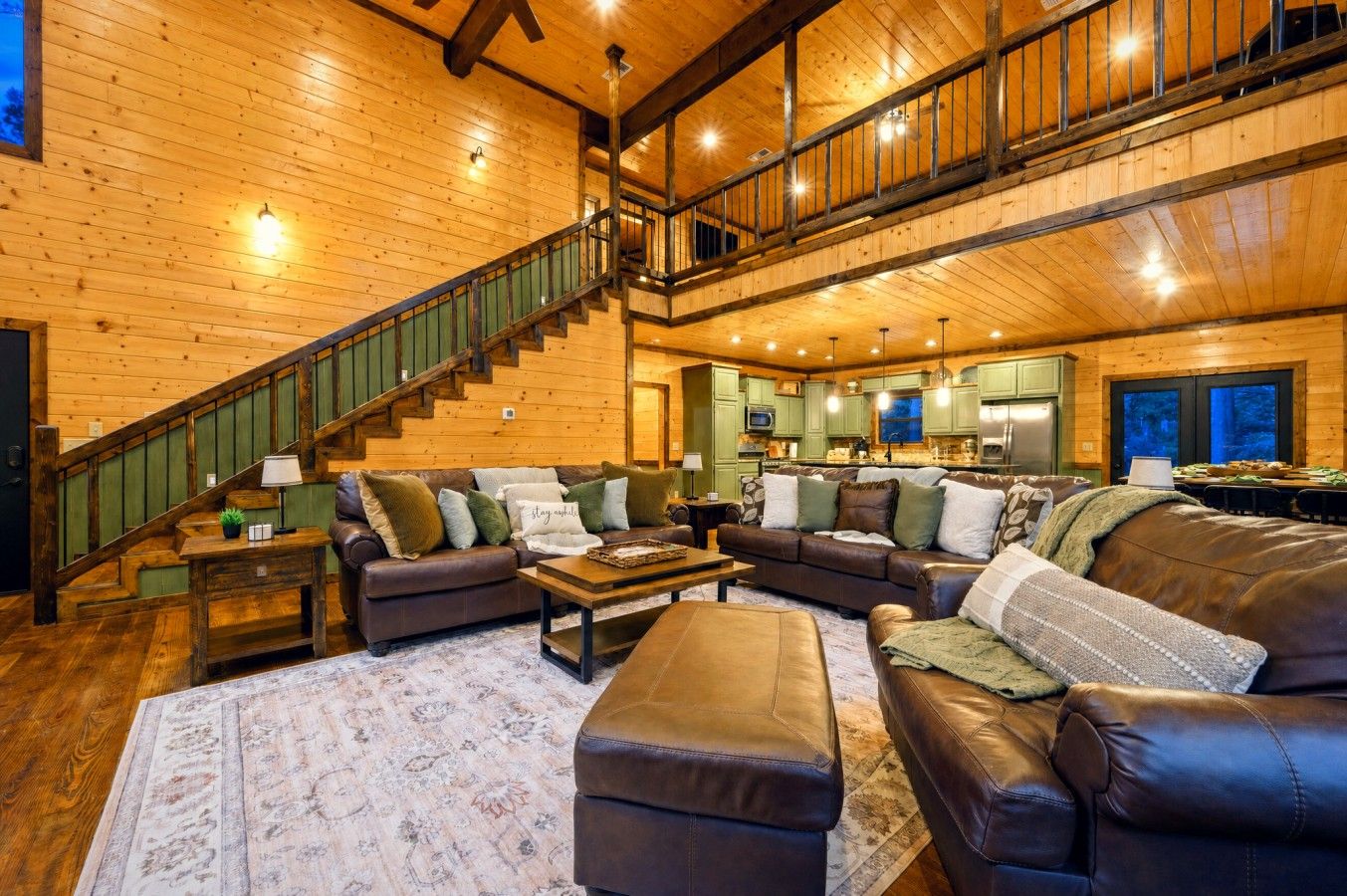 A spacious living area with a high ceiling, wooden walls, a staircase, and a kitchen in the background.
