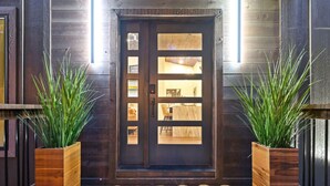 A wooden front door with glass panels, flanked by two wooden planters with green plants, and a mat with "hi" on it.