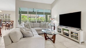 A modern living room with a sectional sofa, a glass coffee table, a flat-screen TV on a wooden stand, and a sliding glass door leading to a balcony.