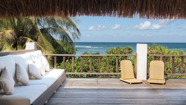 A wooden deck with a white sofa and two wicker chairs, overlooking a beach with palm trees and the ocean. - 4 bedroom Rote Beach House (Rote Barat)