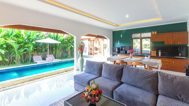 Un salon moderne avec un canapé gris, une table basse en verre agrémentée d’un arrangement floral, et une vue sur une piscine et un espace extérieur.