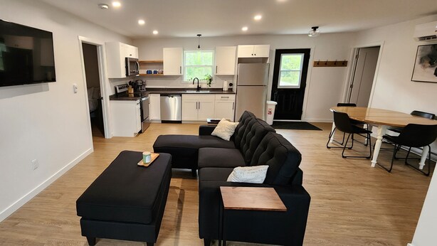 A modern apartment with a living area featuring a black sectional sofa, a small wooden coffee table, and a flat-screen TV mounted on the wall.