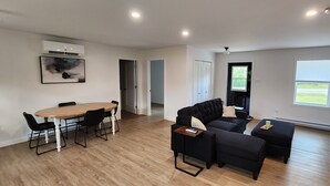 A spacious living room with a wooden dining table, black sofas, and a large window offering a view of the outdoors.