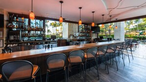 A bar area with a long counter, multiple hanging pendant lights, and a view of palm trees outside.
