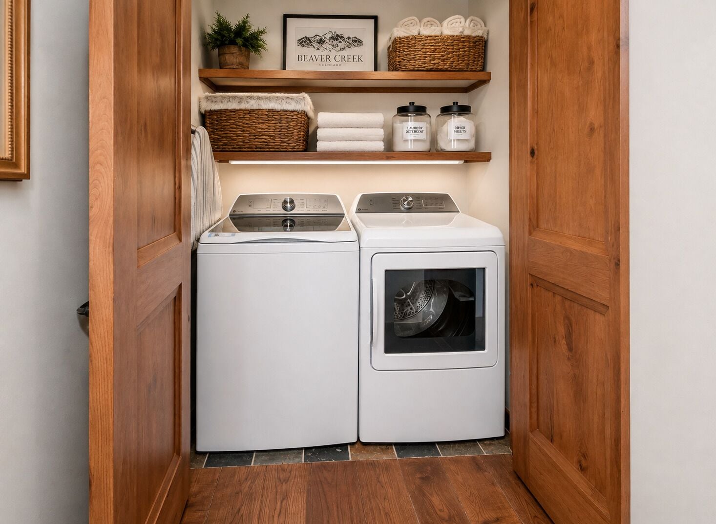 A laundry room with a front-loading washer and dryer, wooden shelving above with baskets and laundry supplies, and a framed sign on the wall.