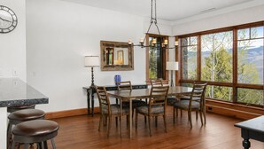 A dining area with a wooden table and chairs, a chandelier, and a view through large windows. - Hummingbird by Elevation Accommodations (Avon)