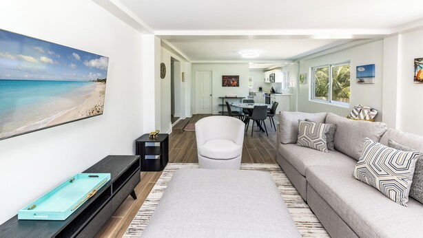 A modern living room with a large flat-screen TV, a grey sofa with patterned cushions, a white armchair, and a wooden coffee table.