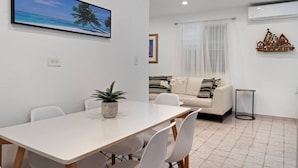 A modern living room with a dining area, featuring a white sofa, a dining table with chairs, a potted plant, and a framed picture on the wall.