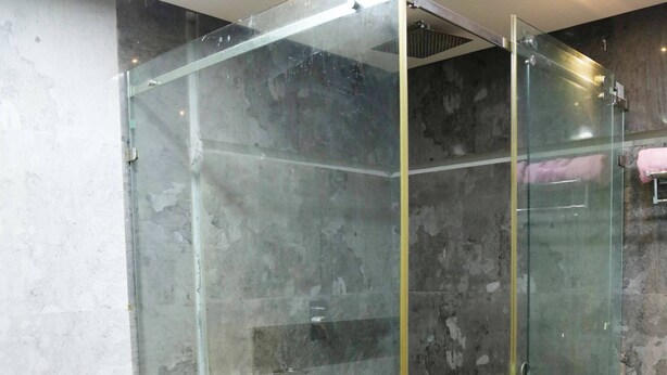 A glass-enclosed shower with a toilet inside, a blue container on the floor, and a patterned rug.