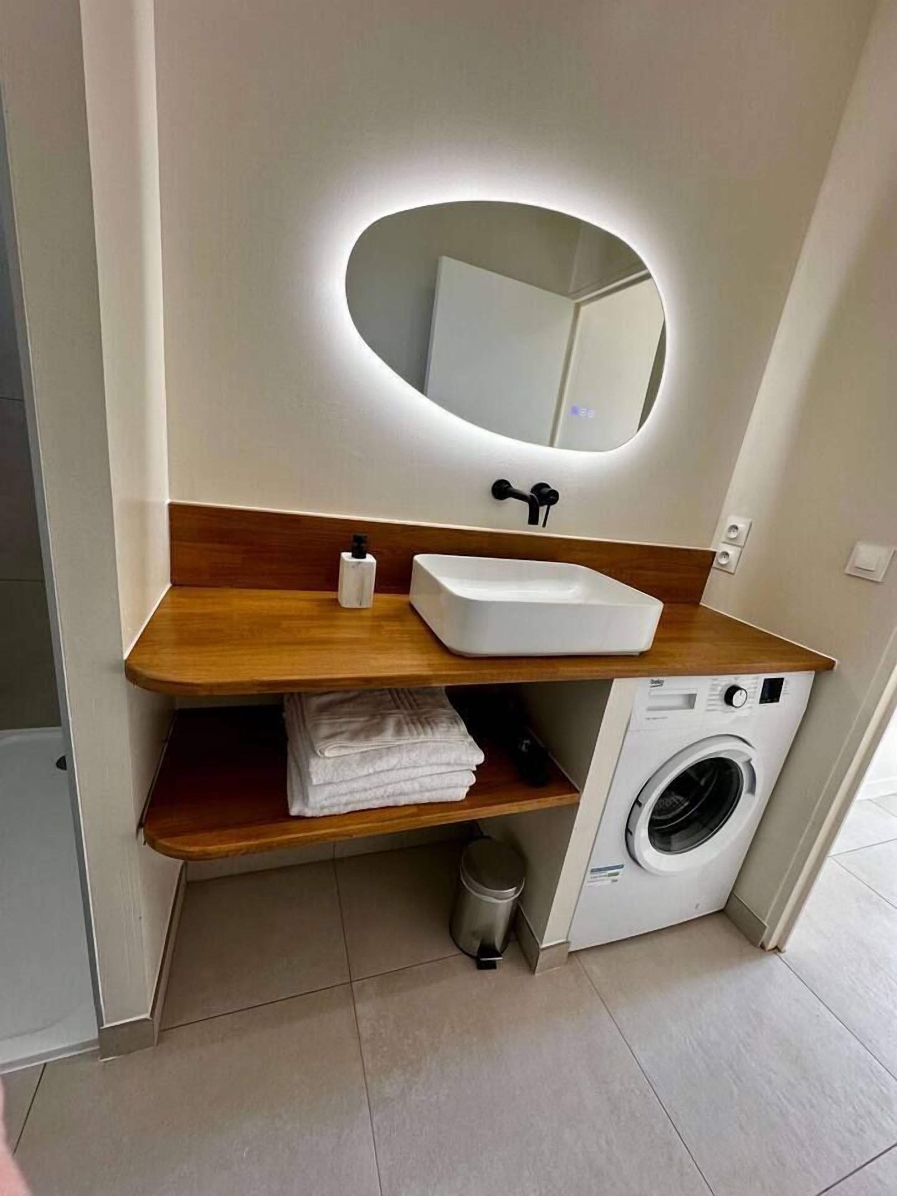 Une salle de bain moderne avec un lavabo blanc, un plan de travail en bois, un miroir encastré et un lave-linge.