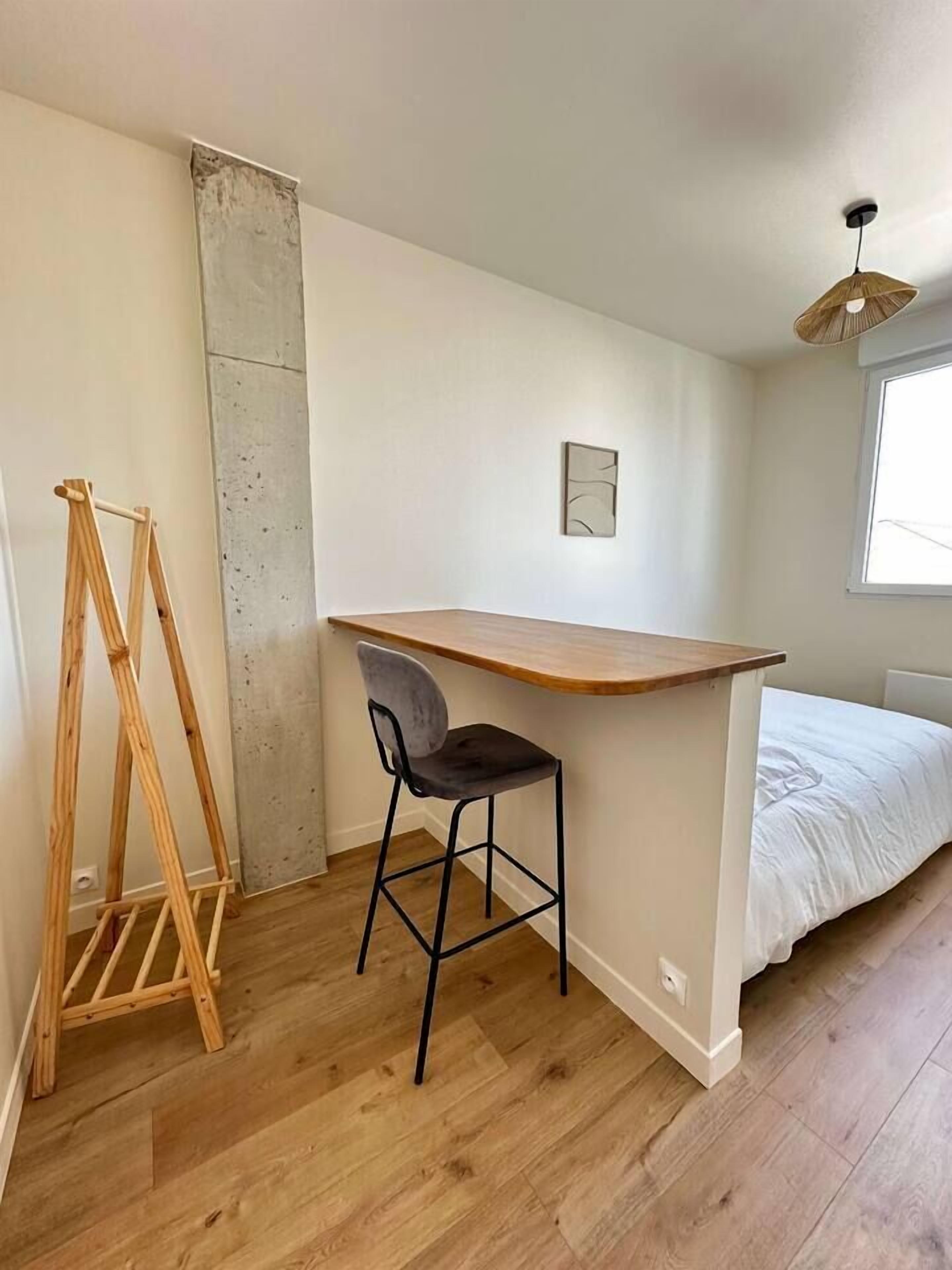 Une petite pièce fonctionnelle, avec un bureau en bois, une chaise noire, un lit avec une literie blanche, une seule œuvre d’art accrochée au mur et une fenêtre laissant entrer la lumière naturelle.