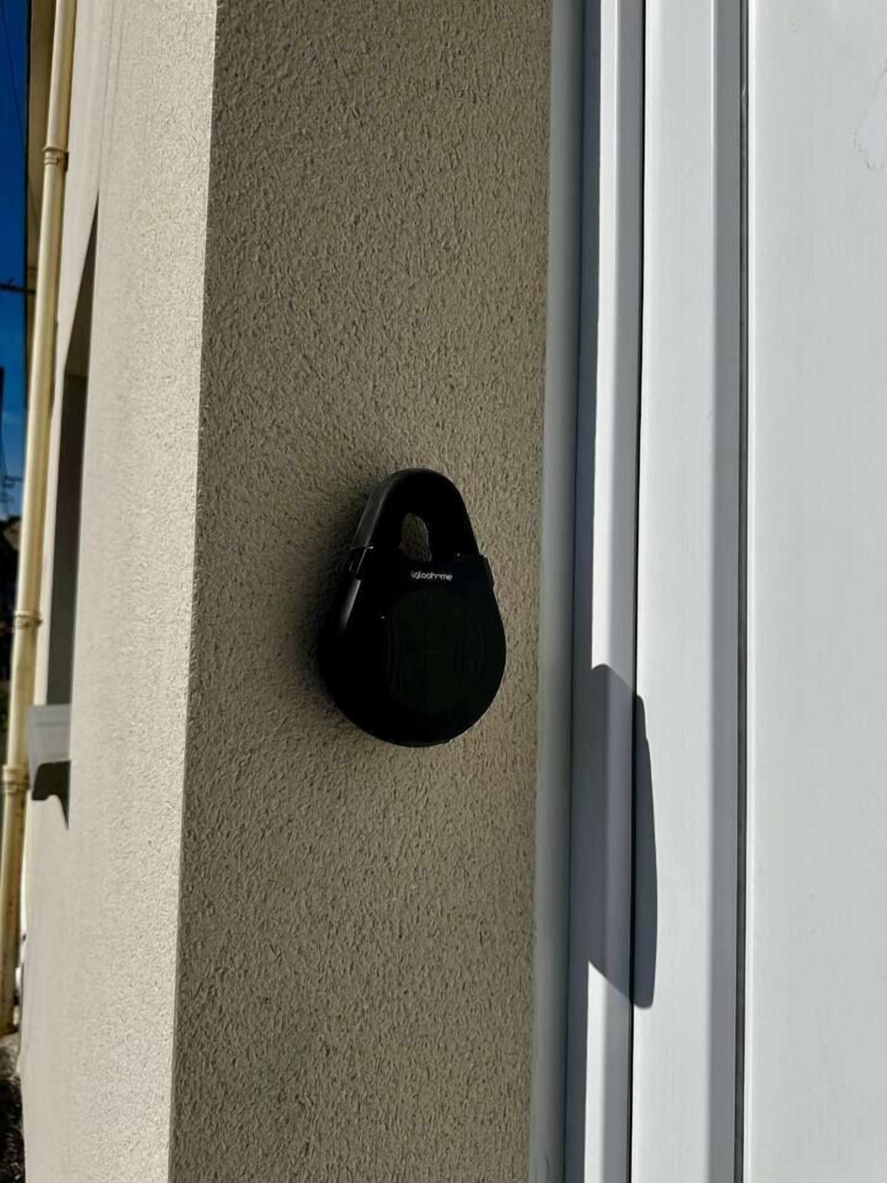 Une sonnette noire est fixée sur un mur beige à texture, à côté d’une porte blanche.
