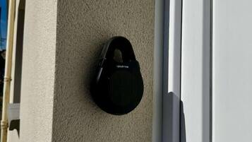 Une sonnette noire est fixée sur un mur beige à texture, à côté d’une porte blanche.