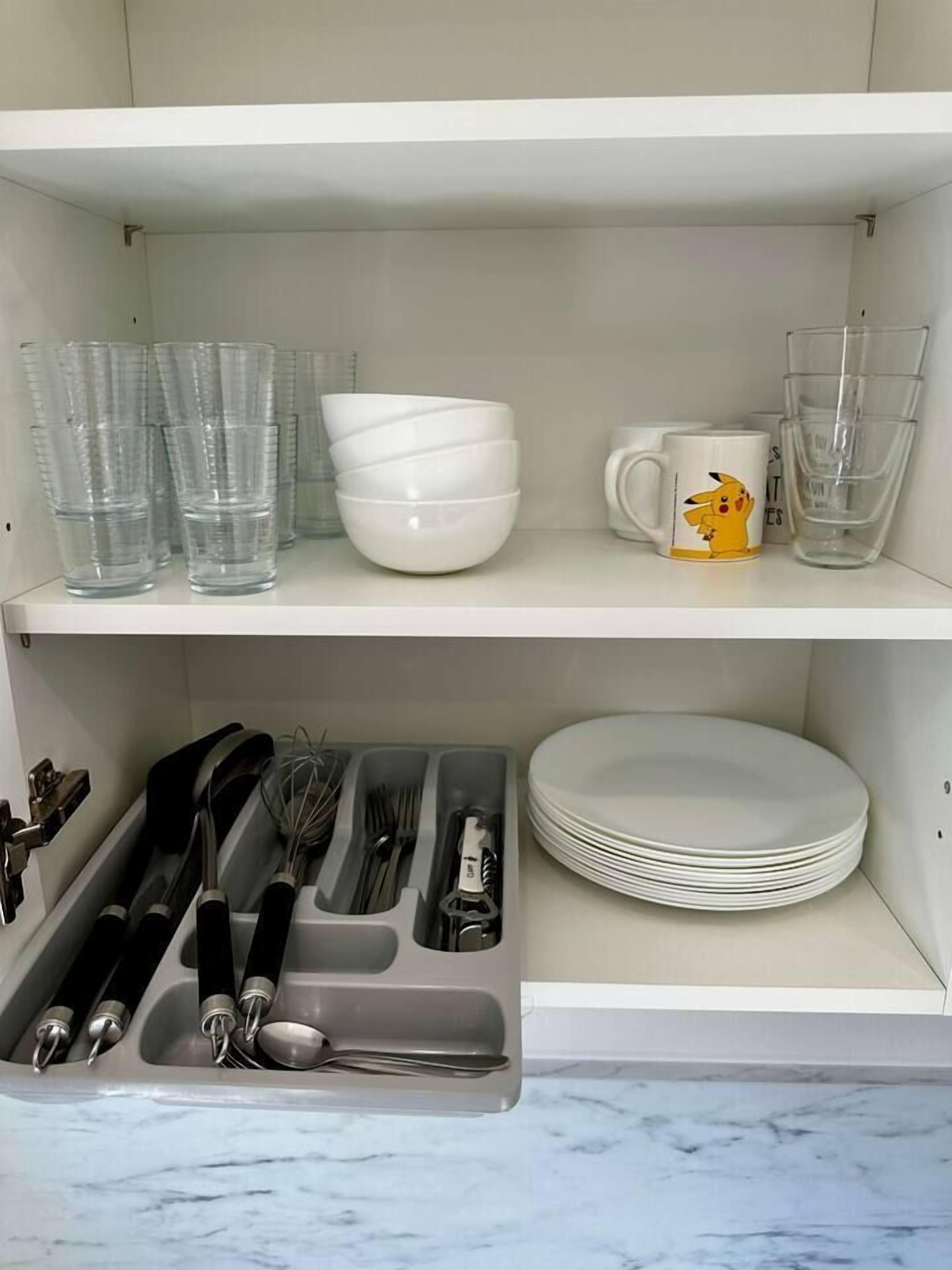 A kitchen with white shelves holding clear drinking glasses, white bowls, and a Pikachu mug. Below, there are utensils and plates in a drawer.