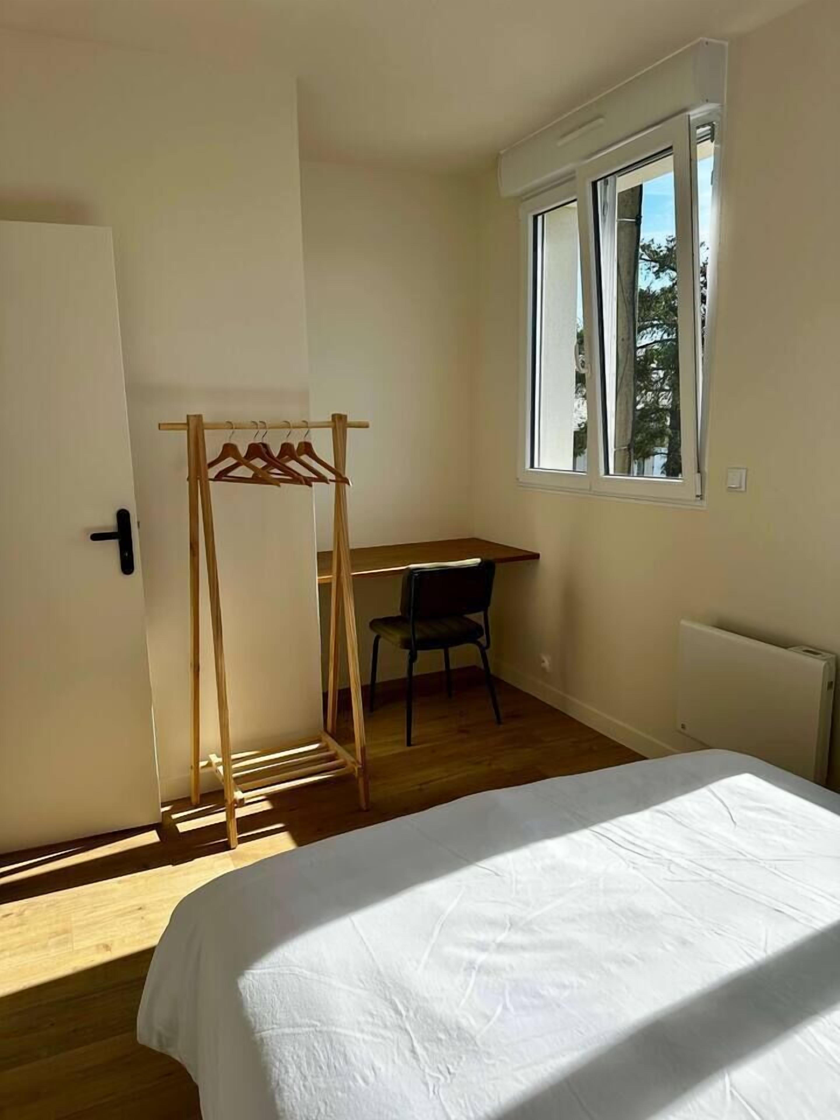 A bedroom with a bed, a wooden chair, a desk, and a clothing rack.