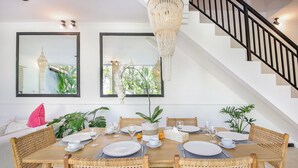 A dining area with a wooden table set for four, surrounded by wicker chairs, with a staircase and a chandelier in the background. - Villa Mandari (Seminyak)