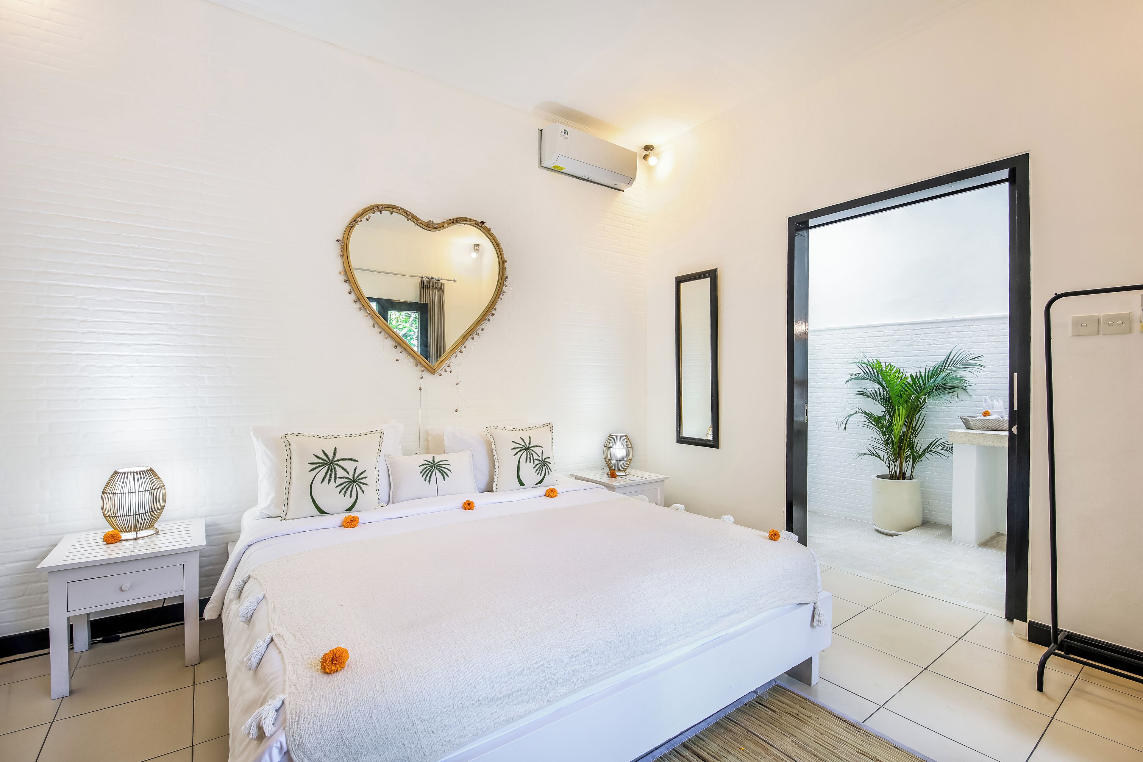 A hotel room with a large bed, a heart-shaped mirror, a small table with a lamp, and a view of a balcony with plants.