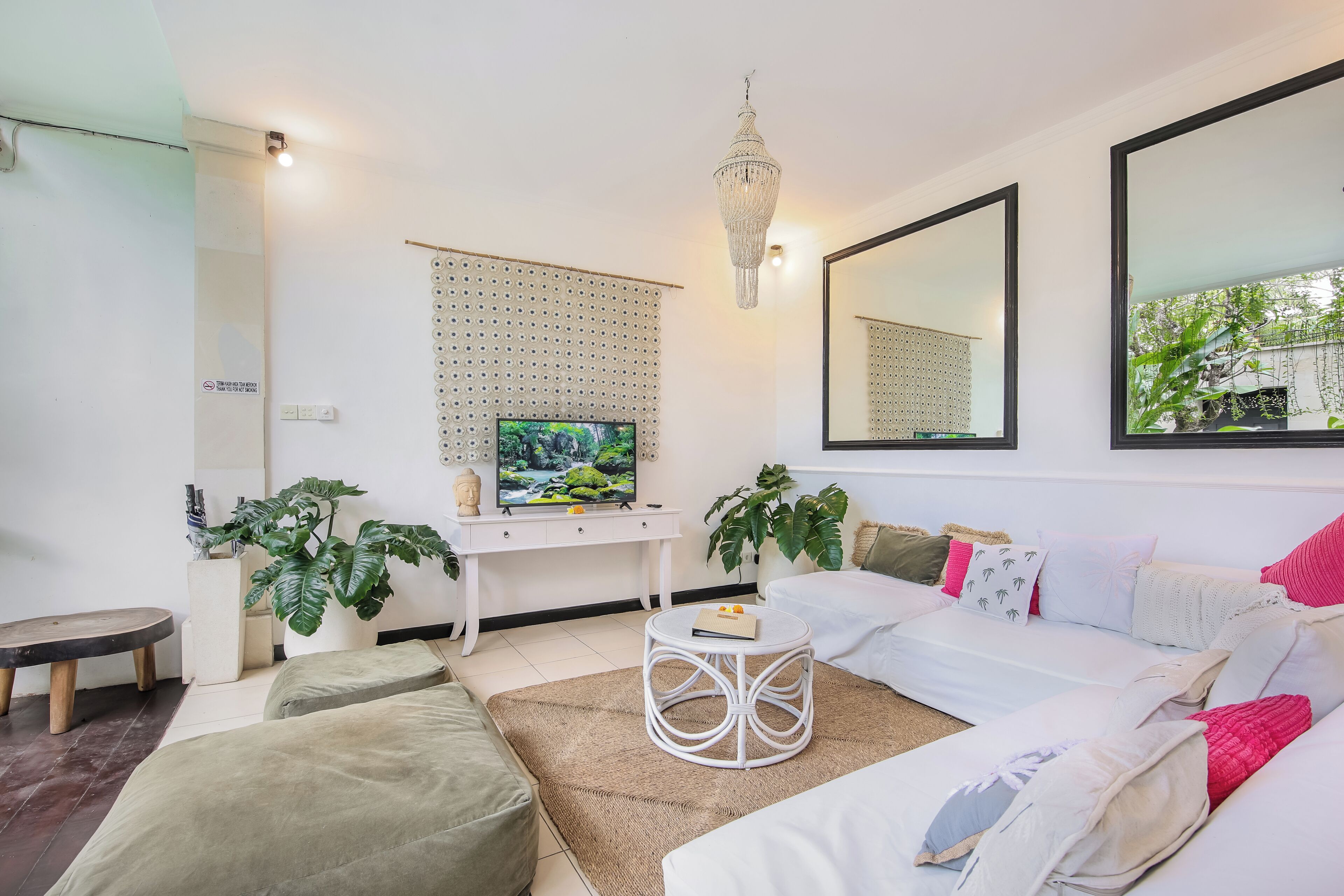 Een moderne woonkamer met een witte bank, een ronde salontafel en een televisie op een witte kast. Twee grote spiegels hangen aan de muur en een kroonluchter aan het plafond.