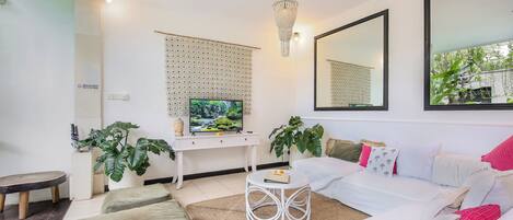 Een moderne woonkamer met een witte bank, een ronde salontafel en een televisie op een witte kast. Twee grote spiegels hangen aan de muur en een kroonluchter aan het plafond.
