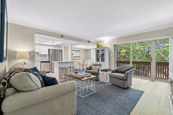 A modern living room with a sofa, armchair, coffee table, and a view of the kitchen and balcony.