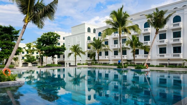 A swimming pool with clear blue water, surrounded by palm trees and a white building with balconies. - Gloca Phu Quoc (Duong To)