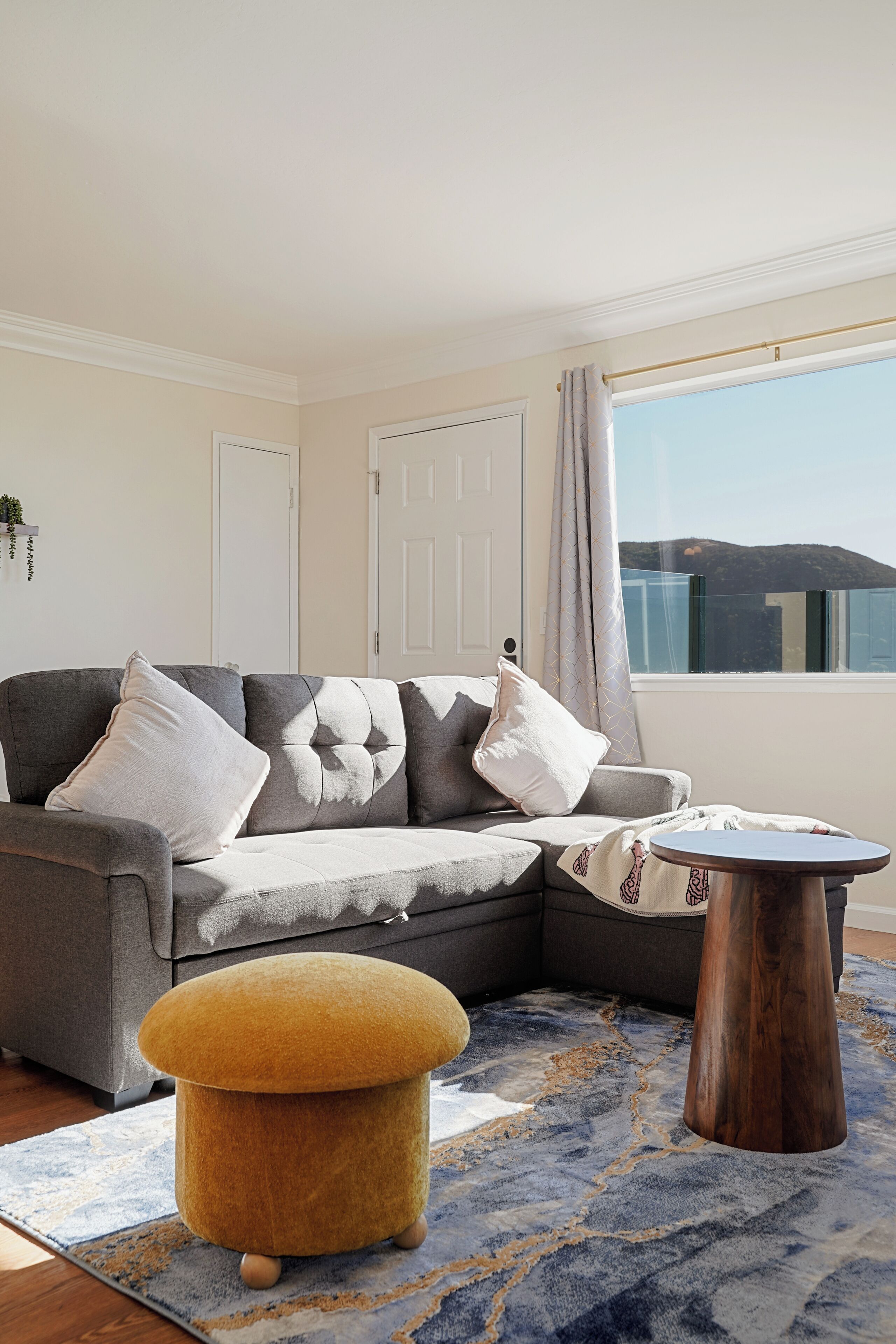 Un salon moderne avec un canapé d’angle gris, une table basse ronde en bois et un pouf de couleur moutarde.